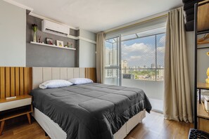 A modern bedroom with a large bed, a bedside table, a shelf with decorative items, and a view of the city through a large window. - Charming Apartment in Pinheiros - Cot0081 (São Paulo)