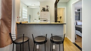 A compact kitchen with a refrigerator, coffee maker, and blender, a bar area with stools, and a bedroom visible through an open door. - Charming Apartment in Pinheiros - Cot0081 (São Paulo)