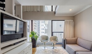 A modern living room with a flat-screen TV, two stylish chairs, a sofa, a large potted plant, and a view of a cityscape through large windows.