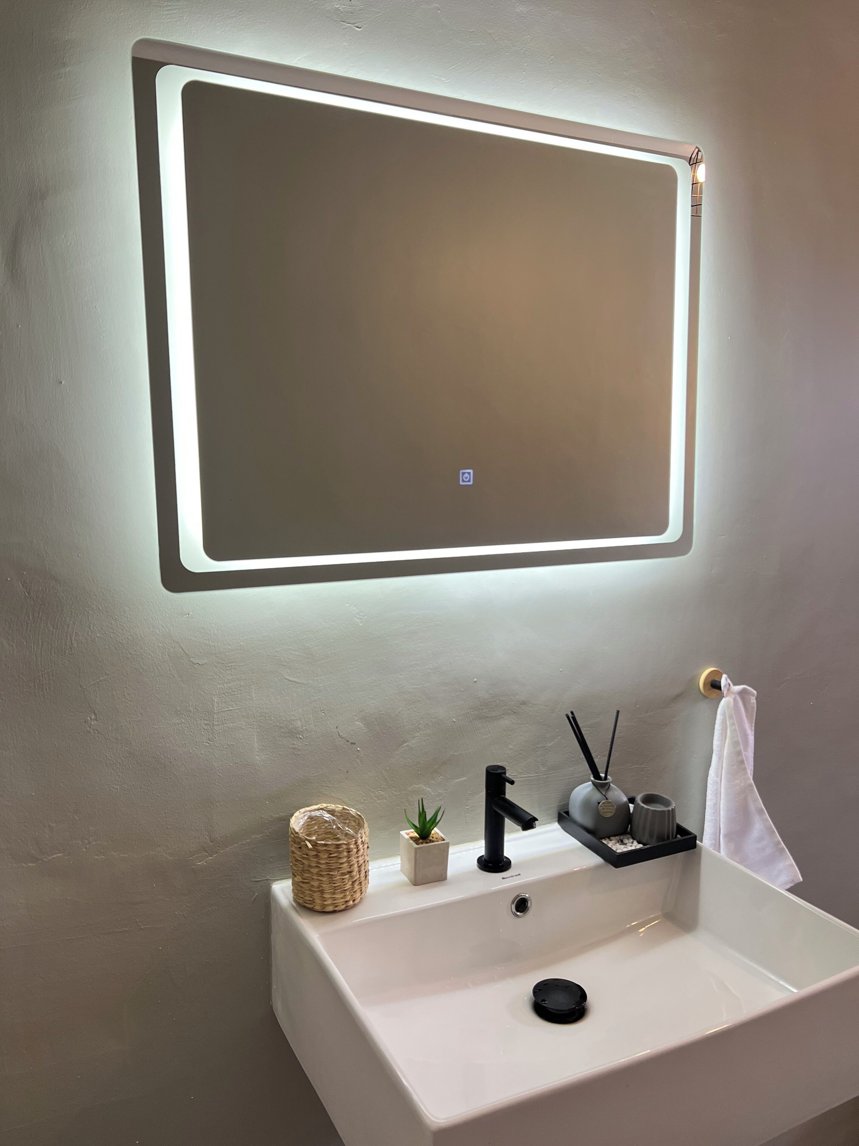 A bathroom with a white sink, a mirror with integrated lighting, a small potted plant, a reed diffuser, and a towel hanging on a hook.