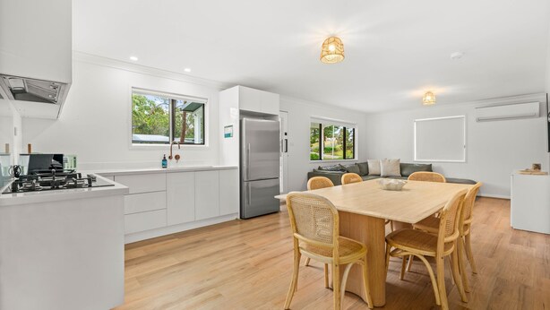Une cuisine moderne avec des meubles blancs, un réfrigérateur en acier inoxydable, un coin repas avec une table en bois et des chaises en osier, et de grandes fenêtres offrant une vue sur la verdure.