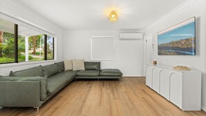 Un salon moderne avec un canapé d’angle, une grande fenêtre donnant sur un paysage verdoyant, un meuble blanc et un tableau accroché au mur.
