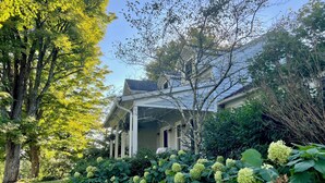 Una casa con porche cubierto, rodeada de exuberante follaje y arbustos de hortensia.