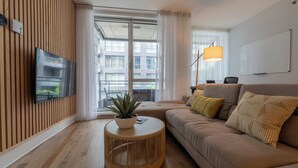 Ein modernes Wohnzimmer mit großem Flachbildfernseher an einer Holzwand, einem runden Couchtisch aus Bambus mit Topfpflanze, einem beigefarbenen Sofa mit dekorativen Kissen und einem großen Fenster mit transparenten Vorhängen.