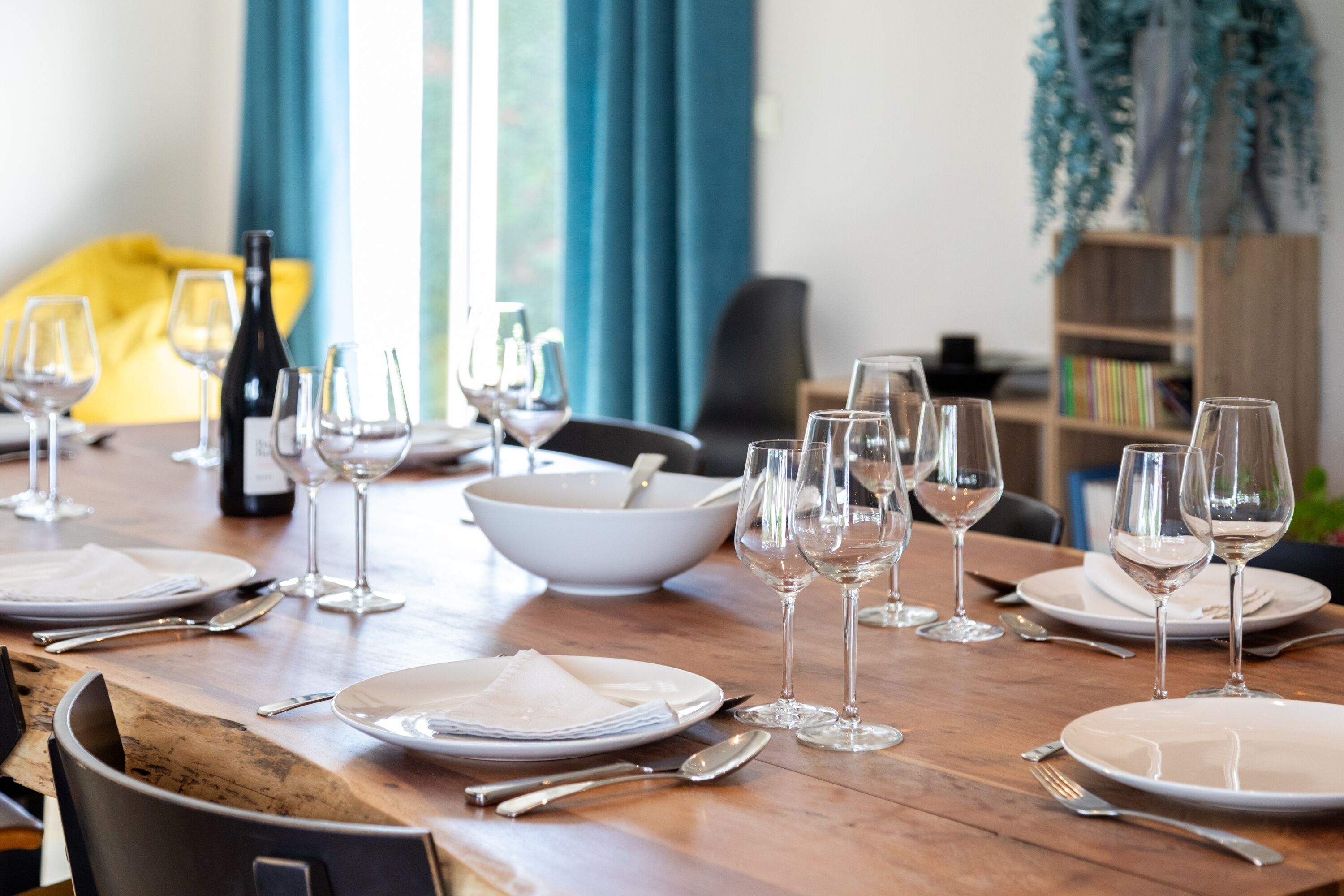 A dining table set with plates, glasses, and cutlery, with a bowl and a bottle in the background.