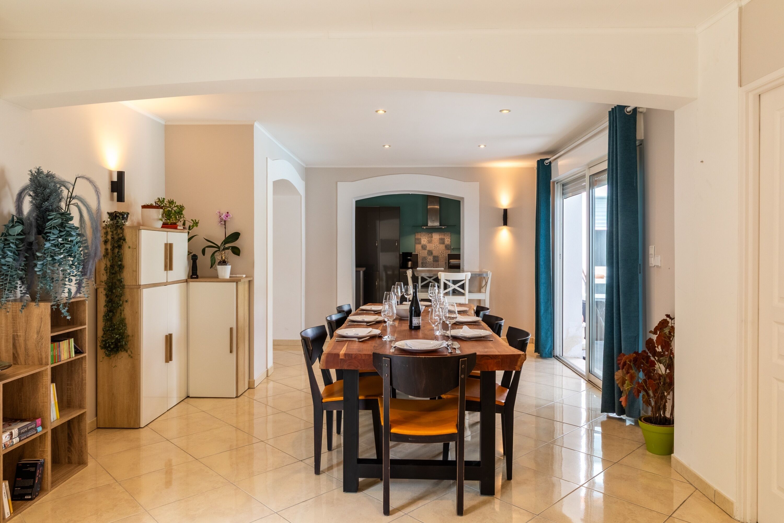 A modern dining area with a wooden table set for six, surrounded by chairs. There is a white cabinet with a plant on top and a bookshelf to the left. The room has a tiled floor and a large window with curtains.