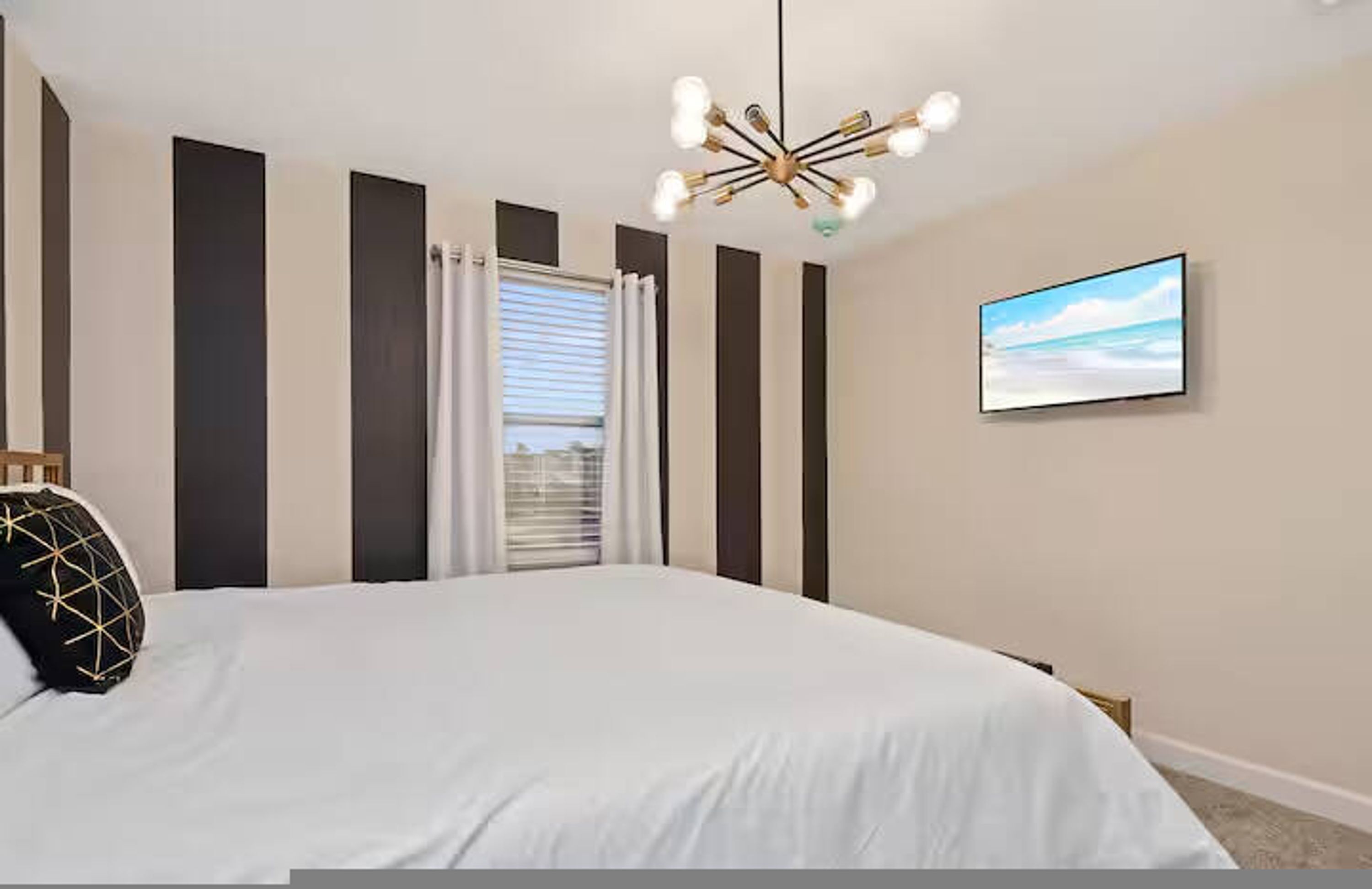 A bedroom with a large bed, a modern chandelier, a window with curtains, and a wall-mounted TV.