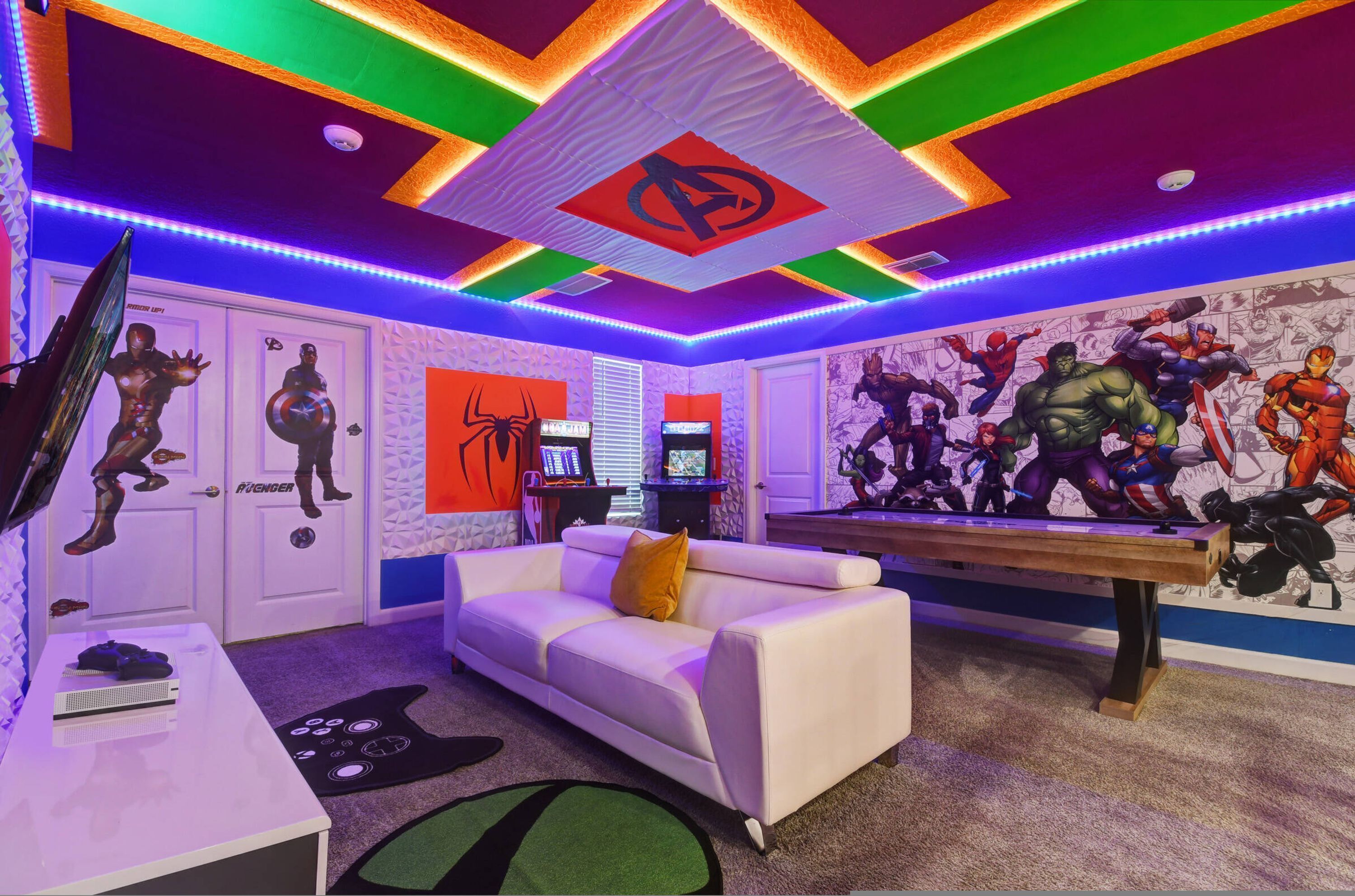 A room with a white couch, a gaming table, and a wall mural of superheroes.