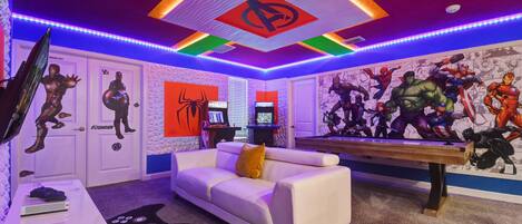 A room with a white couch, a gaming table, and a wall mural of superheroes.