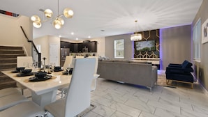 A modern kitchen with dark cabinets and a central island, a dining area with a white table and chairs, and a living area with a grey sofa and blue chairs. - Gorgeous 9bedroom Home Private Pool Game Theater (Kissimmee)