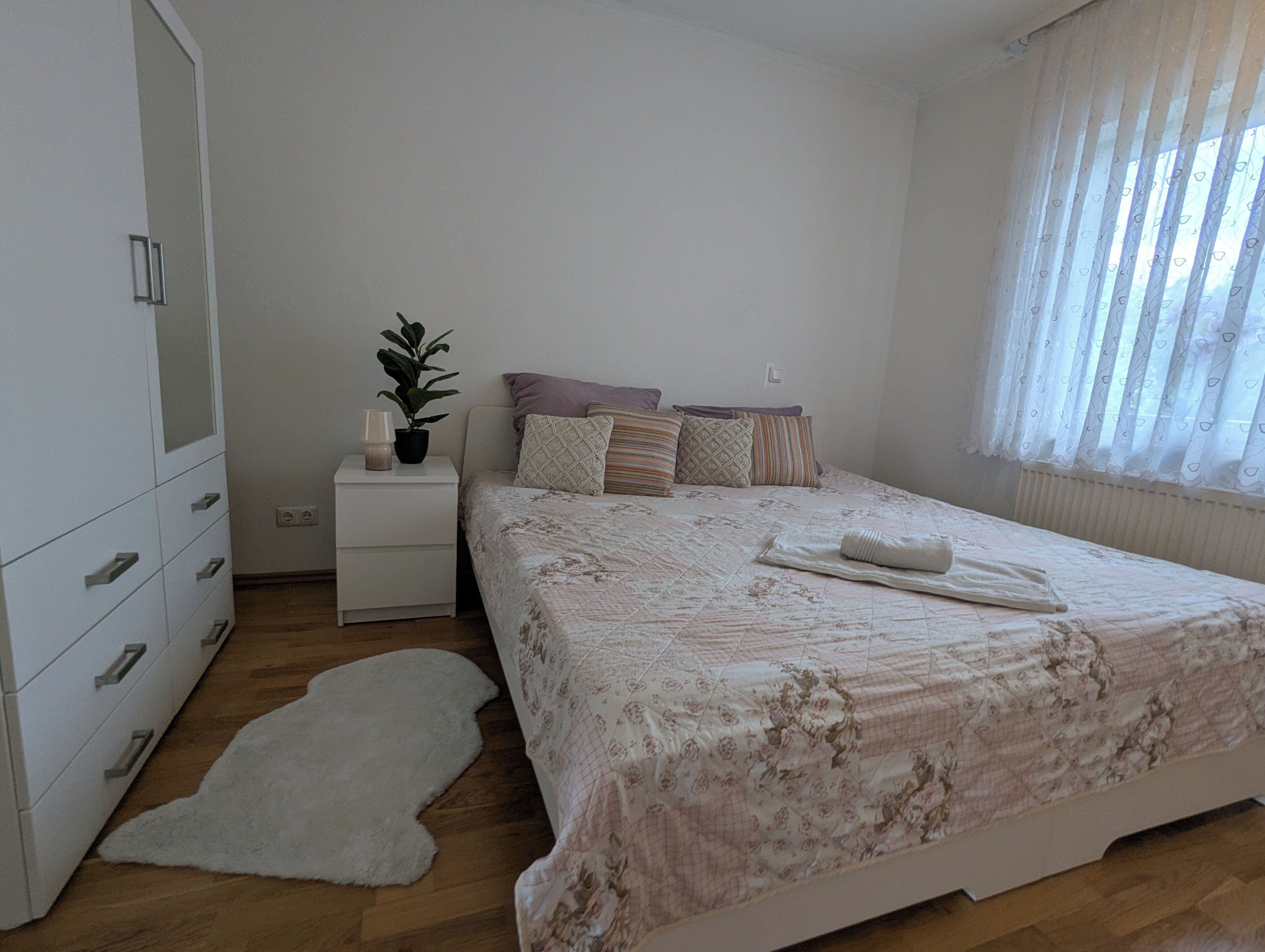 Un dormitori amb llit, una nit, una commode, una planta en test i una finestra amb cortines.