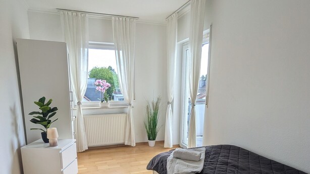 Quarto com cama, mesa de cabeceira, planta em vaso, radiador e janela com cortinas.