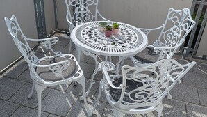 Varanda com mesa redonda e quatro cadeiras brancas, um pequeno vaso de planta e guarda-corpo de metal.