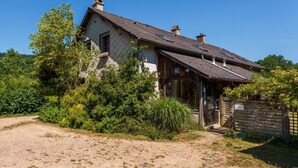 Une maison de campagne au toit en pente, entourée d’une végétation luxuriante et bordée d’un chemin de gravier.