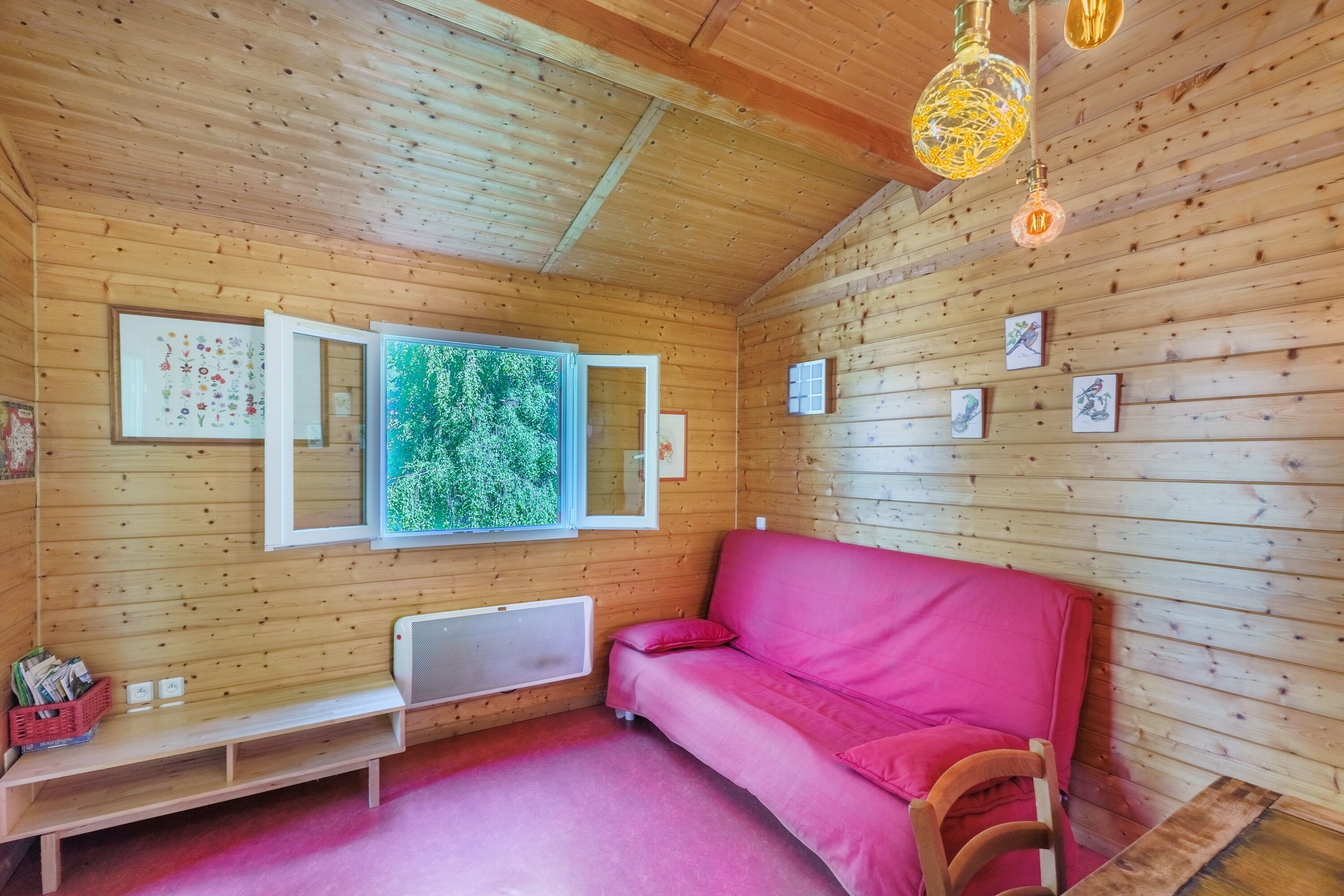 Ein gemütliches Blockhaus mit Holzwänden, einem roten Sofa, einer Bank und einem großen Fenster mit Blick ins Grüne.