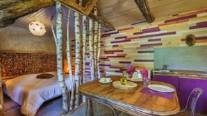 A room with a bed, a dining table with chairs, a wooden ceiling, and a wall with a multicolored pattern.