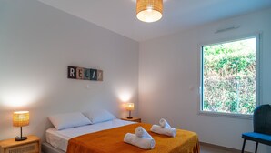 Ein ordentlich bezogenes Bett mit weißen Leinen, ein Nachttisch aus Holz mit einer Lampe und ein Fenster mit Blick auf Grünflächen.
