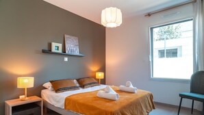 A neatly arranged bedroom with a bed, bedside lamps, a chair, a window, and a wall-mounted shelf with a picture. - Maison HG - Presqu'ile de Crozon (Plomodiern)