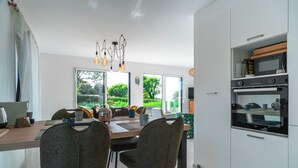 A modern kitchen with white cabinets, a built-in oven, and a dining area with a wooden table and two chairs. - Maison HG - Presqu'ile de Crozon (Plomodiern)