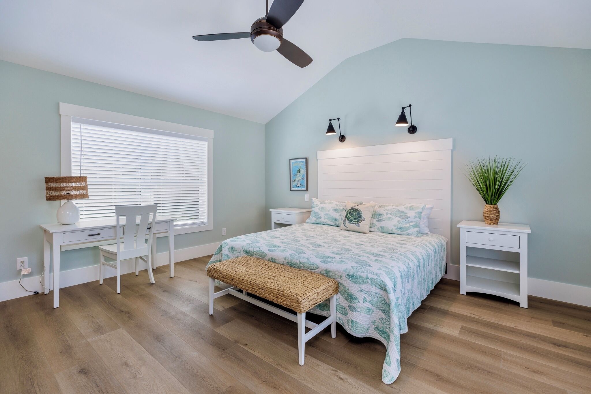 Une chambre à coucher comprenant un lit, un bureau, une chaise, un banc, un ventilateur de plafond, des lampes de chevet et une table de chevet agrémentée d’une plante.