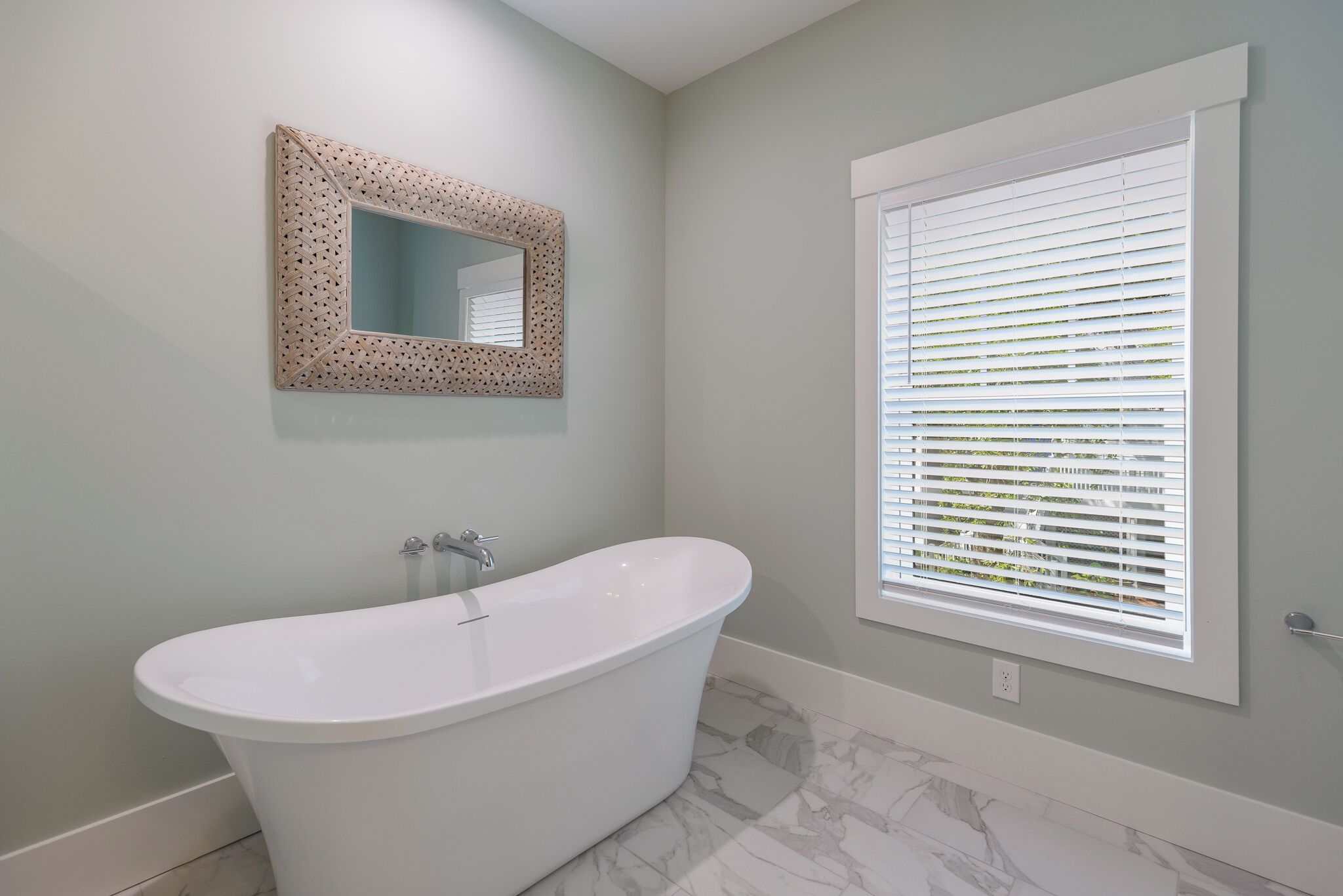 Un baño con una bañera independiente, un espejo instalado en la pared con un marco decorativo y una ventana con persianas.