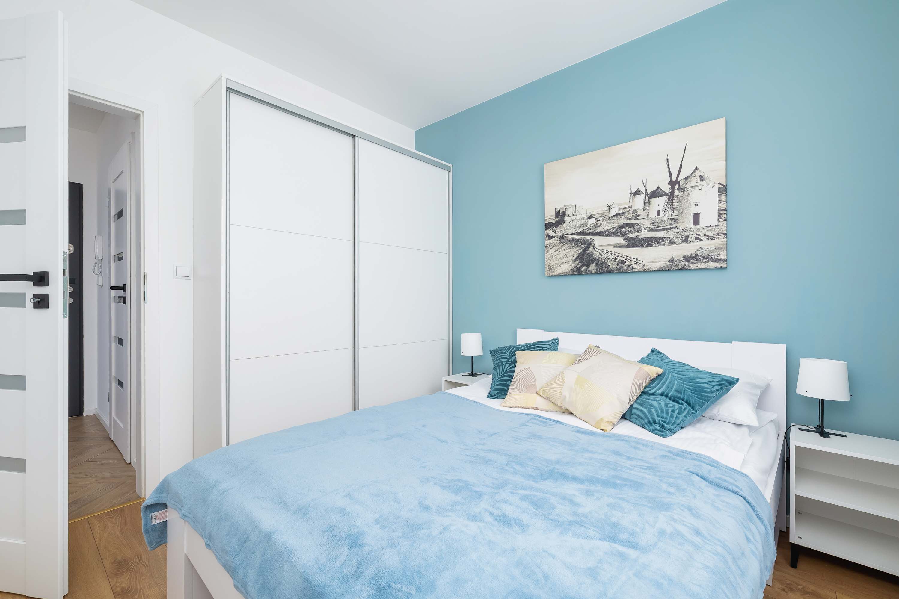 A bedroom with a bed, a nightstand, a bedside table, a wardrobe, and a wall-mounted picture.