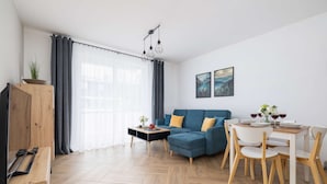 A modern living room with a sofa, dining table, and chairs. There is a television, a plant, and artwork on the walls.