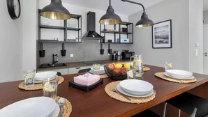 A modern kitchen with a wooden dining table set for four, equipped with black and gold pendant lights, a black clock, and a framed landscape picture on the wall. - Elegant Apartment | 150 m from the Beach, Terrace, Parking (Międzywodzie)