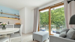Una sala de estar moderna con un sofá gris, una mesa de comedor blanca y un balcón con vistas a árboles.