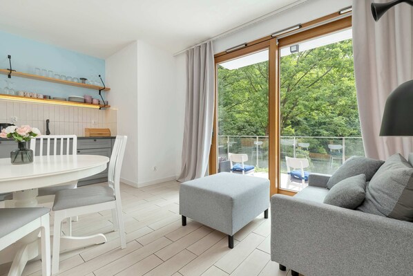 A modern living room with a grey sofa, a white dining table, and a balcony with a view of trees.