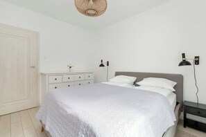 A bedroom with a bed, white walls, a wooden door, a nightstand, and a wall-mounted lamp.