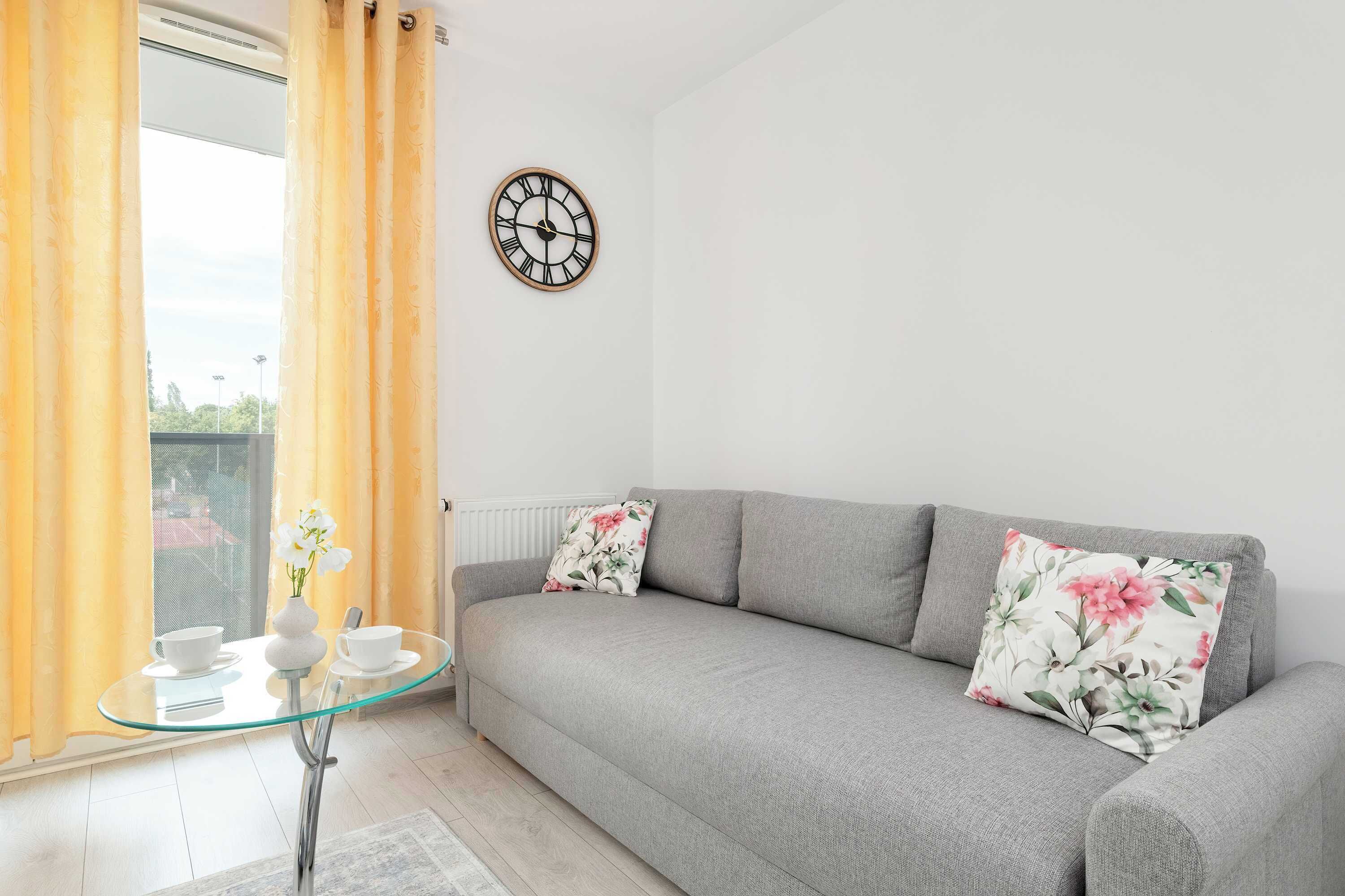 A modern living room with a grey sofa, floral cushions, a glass coffee table, and a wall clock.