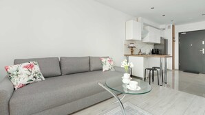 Una sala moderna con un sofá gris, una mesa de centro de vidrio y una cocina al fondo.