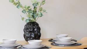 A dining table with white bowls and plates, a black vase with green leaves, and wooden chairs with black cushions. - Cozy Apartment in the Heart of the Old Town | Smart TV and Wi-Fi (Warszawa)