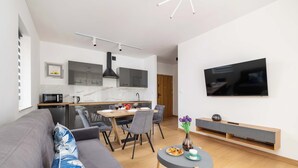 Moderni olohuone, jossa on harmaa sohva, ruokailutila puupöydällä ja -tuoleilla, valkoisilla kaapeilla varustettu keittiö ja musta liesituuletin sekä seinälle asennettu televisio.