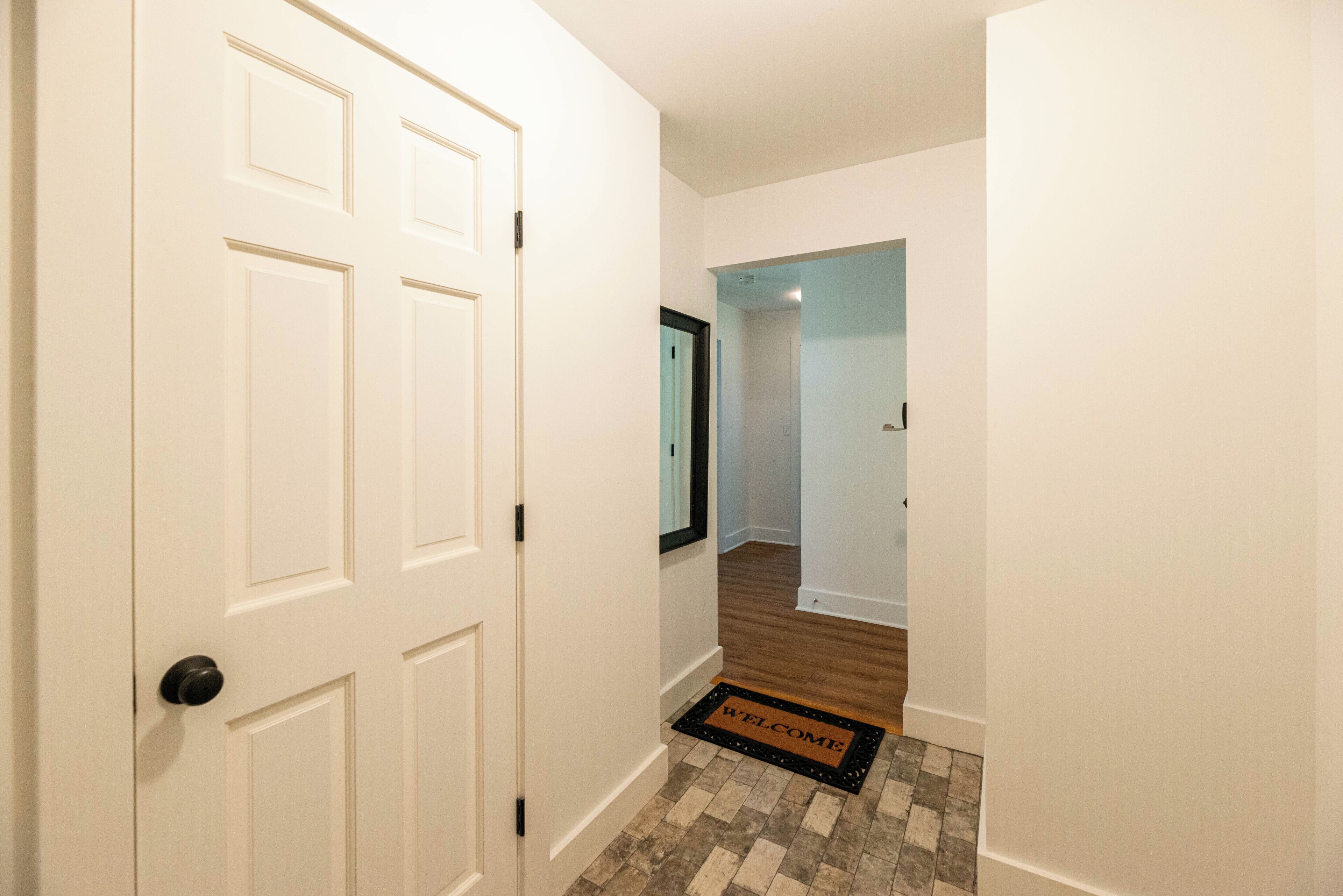 A hallway with a white door, a welcome mat, and a wooden floor.
