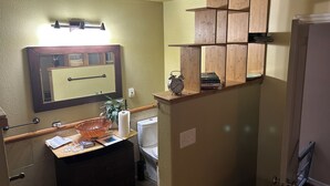 A bathroom with a wooden vanity, a mirror, a shelf with books, and a toilet.