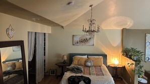 A bedroom with a bed, a chandelier, a bedside table, a mirror, a window with curtains, and a wall-mounted artwork.