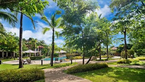 A pathway leading to a pool area surrounded by lush greenery and palm trees, with lounge chairs and umbrellas. - Laguna Village, Two-Bedroom 138/25 Townhouse (Cherngtalay)