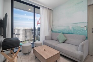 Un salon moderne avec un canapé, un pouf et une table basse, un balcon offrant une vue sur les grues de construction, et un mur orné d’œuvres d’art abstraites.