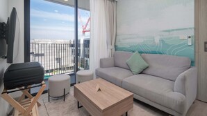 En moderne stue med sofa, skammel og sofabord, en balkon med udsigt til byggekraner, og en væg med abstrakt kunst.