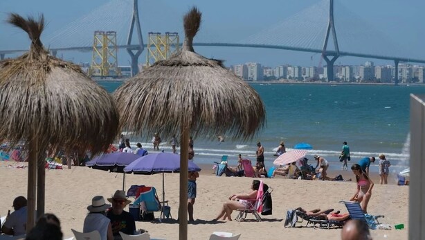 Een strand met rieten parasols, ligbedden en mensen die ontspannen. In de verte is een brug met gele torens te zien.
