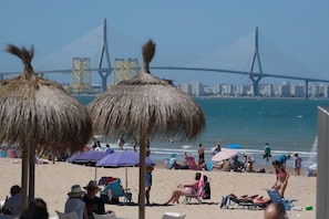 A beach with thatched umbrellas, beach chairs, and people relaxing. A bridge with yellow towers is visible in the background.