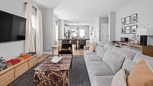 A modern living room with a large flat-screen TV, a grey sofa, a coffee table, and a dining area in the background. - Modern 3-bedroom home with fenced yard + easy parking (Indianapolis)