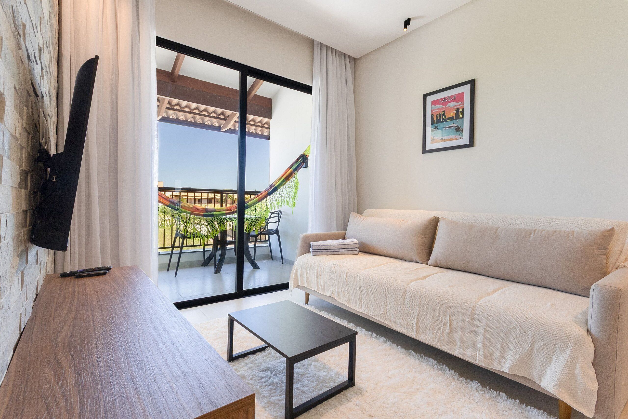 A living room with a sofa, a coffee table, a TV mounted on a stone wall, and a balcony with a hammock.