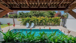 A pool area with a wooden pergola overhead, surrounded by lush greenery and a view of a residential area.