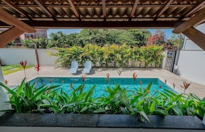 A pool area with a wooden pergola overhead, surrounded by lush greenery and a view of a residential area.