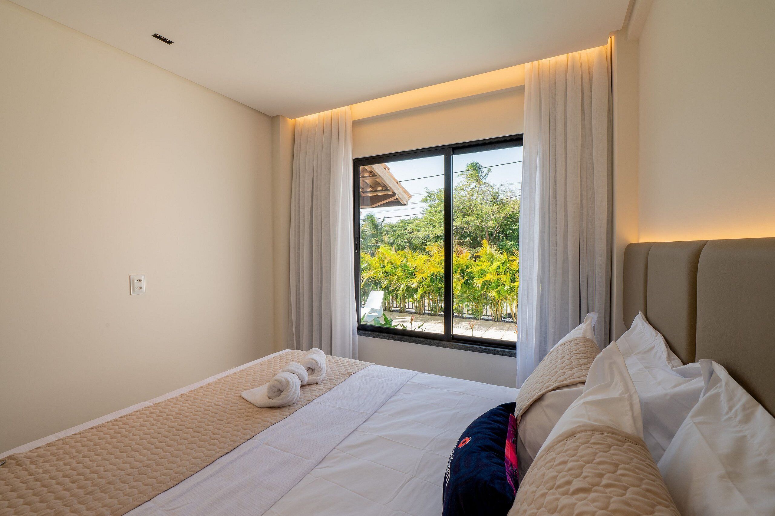 A hotel room with a bed, a window with curtains, and a view of greenery outside.