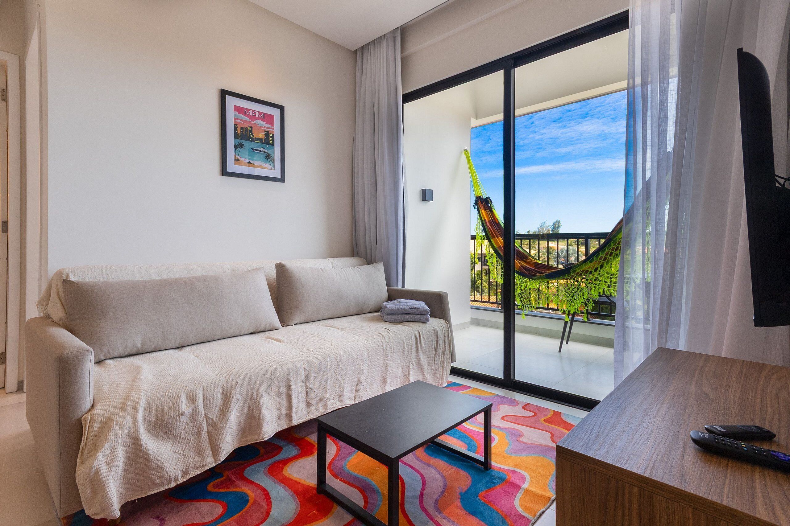 Ein modernes Hotelzimmer mit einer Couch, einem kleinen Tisch, einem farbenfrohen Teppich, einem Balkon mit einer Hängematte und einem großen Fenster.