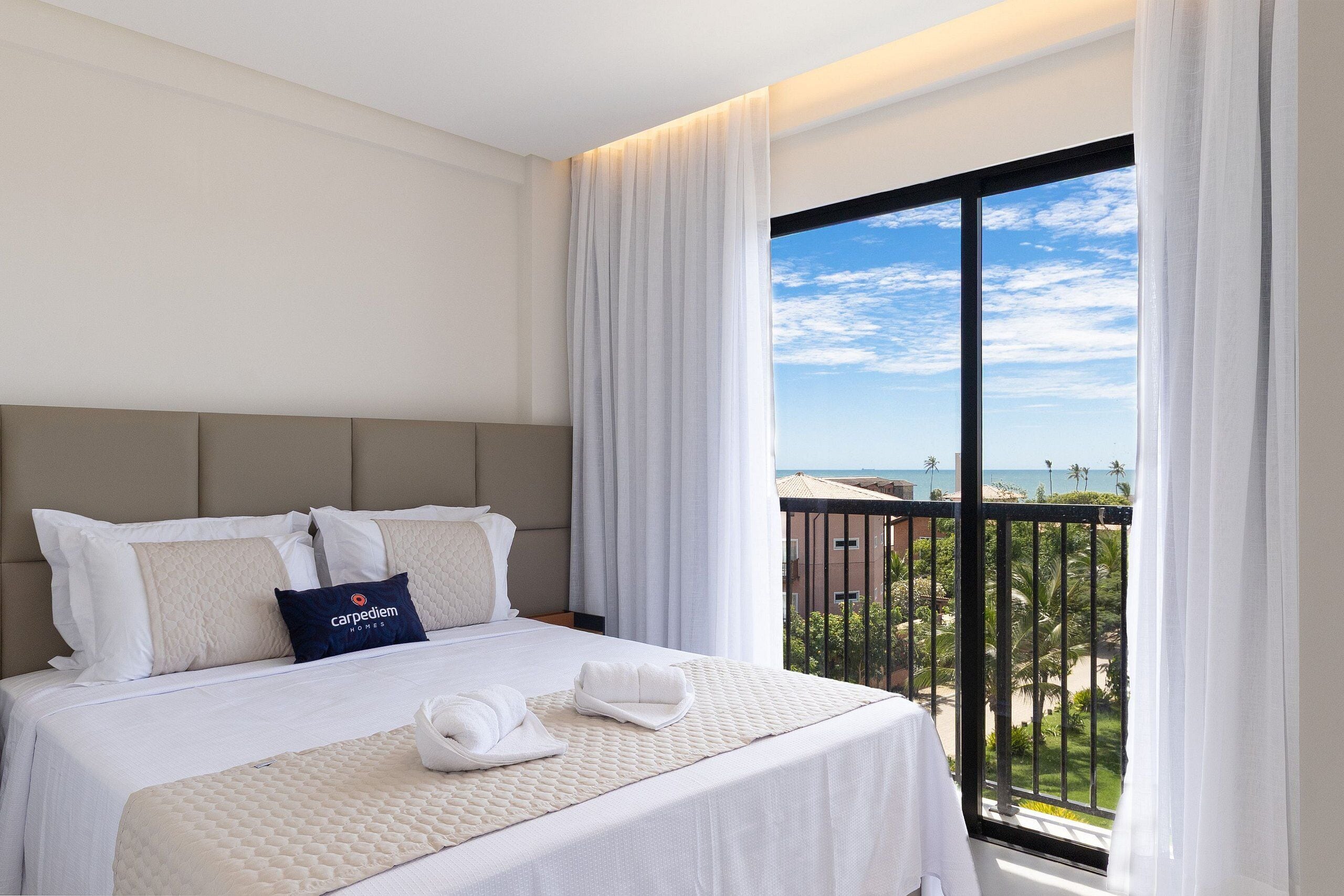 A hotel room with a large bed, white bedding, a headboard, and a view of the beach through a balcony.